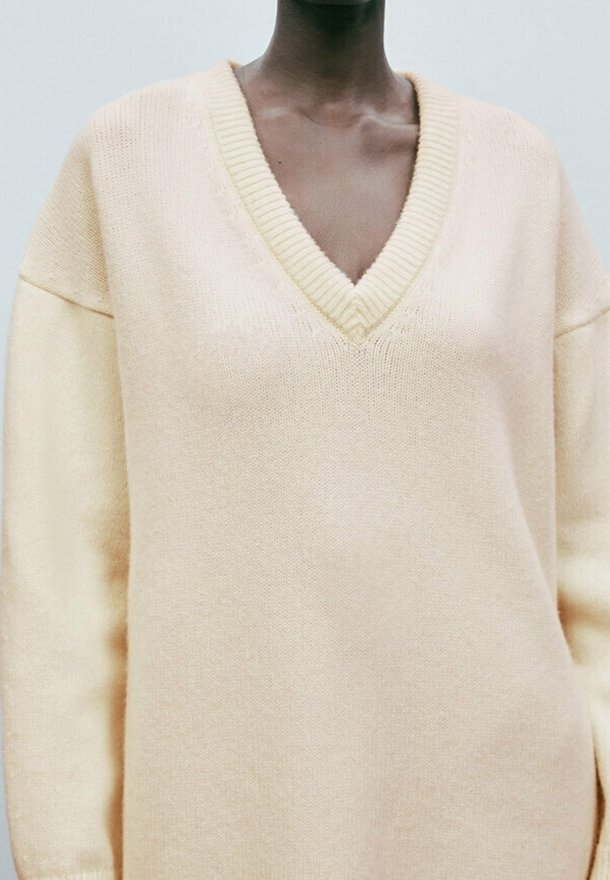 Personne portant un pull en maille oversize couleur crème avec un profond col en V et des manches longues, montré des épaules jusqu'aux hanches.
