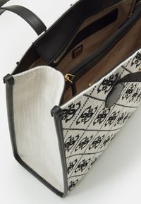 Canvas totebag met een zwart-wit geometrisch patroon, zwarte leren accenten, ritssluitingen en gouden metalen accenten.