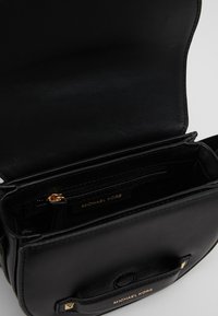 Sac à main en cuir noir avec un intérieur zippé, des accents en métal doré et un logo de marque en relief. Design compact avec plusieurs compartiments.
