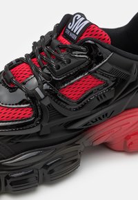 Schwarzer und roter Sportschuh mit glänzendem Obermaterial, Mesh-Einsätzen, dicker Außensohle mit einem Farbverlauf in Rot und Schwarz sowie geschichteten Details.