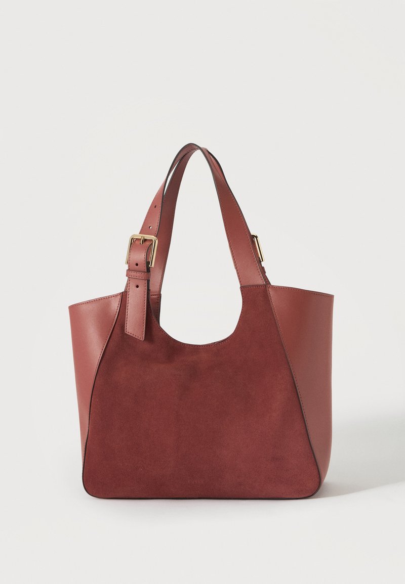 Sac cabas en cuir marron avec panneau avant en daim et bretelles ajustables avec boucles dorées sur fond uni.