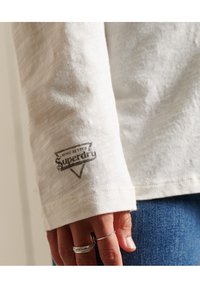 Långärmad krämfärgad bomullsskjorta med en grå logotyp på ärmslutet och synlig struktur. Hand med silverringar visas bredvid skjortan.