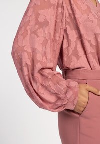 Durchsichtige, pinke Bluse mit floraler Stickerei, voluminösen Ärmeln und glatter Textur, kombiniert mit einer eng geschnittenen pinken Hose mit seitlichem Taschen.