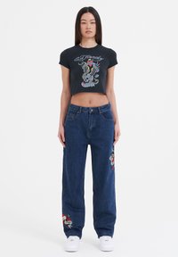 Black cropped t-shirt with graphic print featuring a skull and colorful designs. Loose blue denim jeans with embroidered accents. White sneakers.