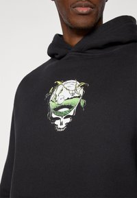 Μαύρο φούτερ με κουκούλα που παρουσιάζει ένα γραφικό σχέδιο με ένα κρανίο και πράσινες λεπτομέρειες, απεικονίζοντας ένα τοπίο μέσα στο περίγραμμα του κρανίου και φυλλώματα αμπέλου.