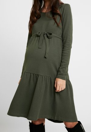 Kobieta w ciąży ubrana w oliwkowozieloną, długorękawową sukienkę ciążową z kokardą zawiązaną nad brzuchem oraz czarne kozaki za kolano.