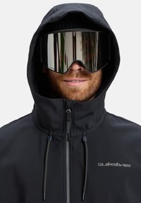 Black jacket with a hood, featuring a front zip and drawstrings. Mirrored ski goggles complete the look, with the brand name on the chest.