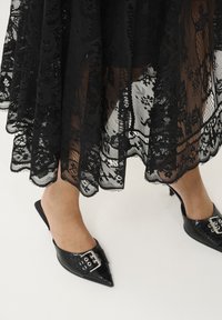 Gonna in pizzo nero con intricate fantasie floreali, design a strati e tessuto semitrasparente, abbinata a mule nere a punta con dettagli in metallo.