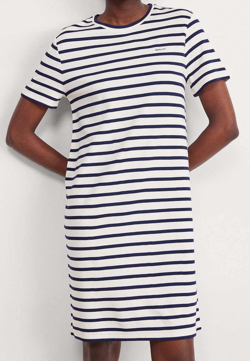Personne portant une robe blanche à manches courtes avec des rayures horizontales bleu marine et un petit logo sur la poitrine.