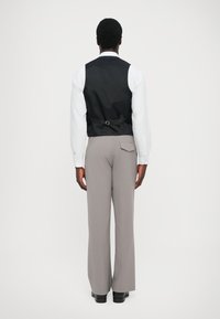 Gilet noir avec un détail de boucle sur une chemise blanche, associé à un pantalon large gris avec une poche arrière. Texture de tissu lisse.