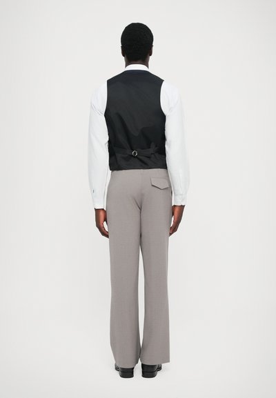 Schwarze Weste mit Rückenriemen, weißes Langarmhemd, graue Bundfaltenhose und schwarze Schuhe. Glattes Material und maßgeschneiderte Silhouette.