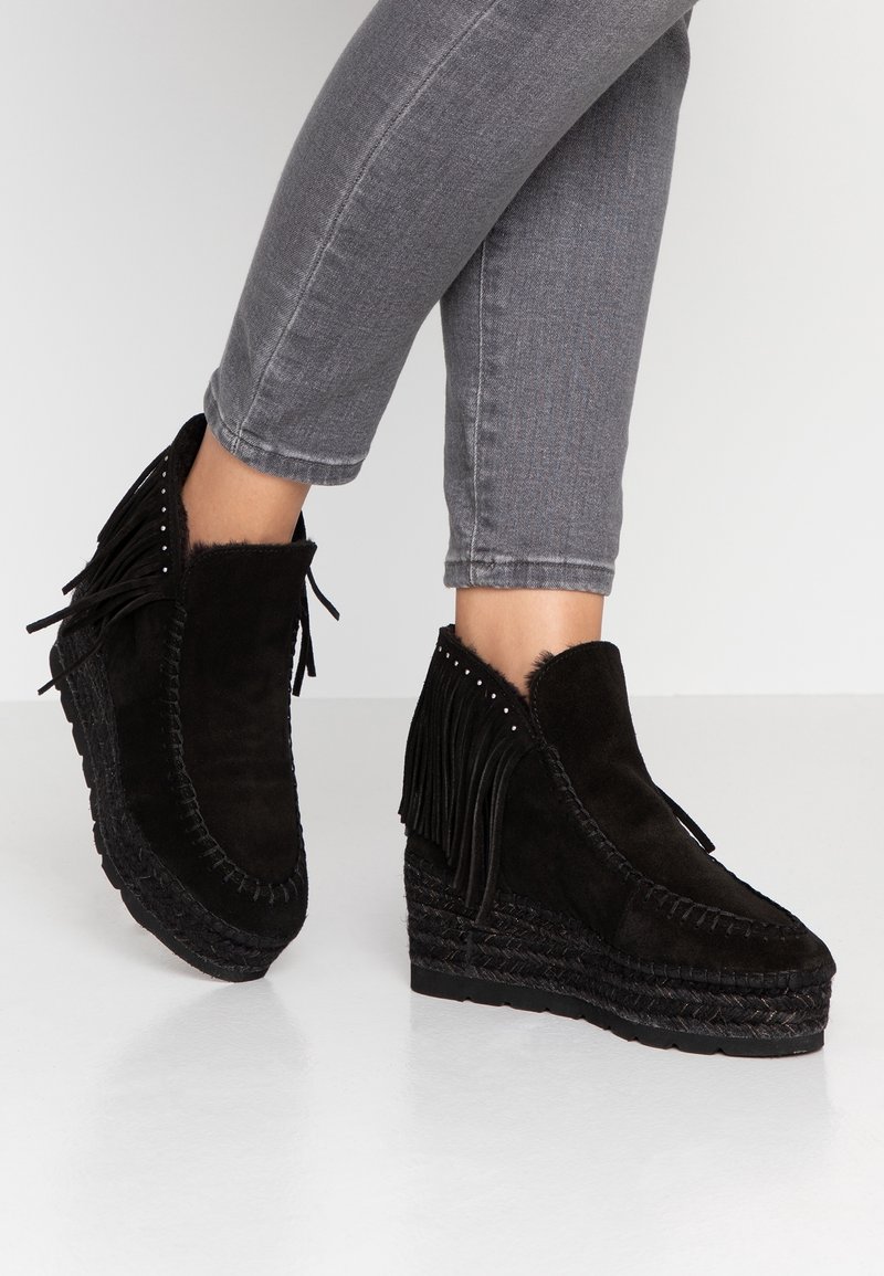 Bottines noires à talons compensés avec des détails en franges portées avec un jean skinny gris sur un fond uni.