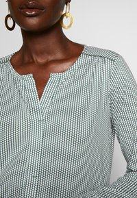 Blusa a pois verde e bianca con scollatura a a V, spalle plissettate e maniche lunghe. Caratterizzata da una texture di tessuto liscia.
