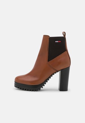 Bottine en cuir marron avec panneau latéral élastique noir, talon haut épais, languette de traction et petite écusson logo rouge, blanc et bleu.
