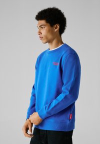 Blauwe katoenen sweatshirt met geribbelde manchetten en zoom. Heeft een klein rood logo op de borst en een zijlabel met merkdetails.
