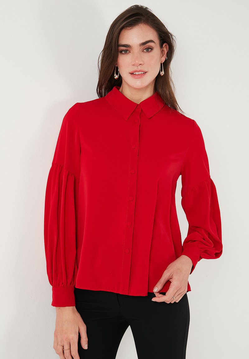 Red, long-sleeve blouse with a button-up front, collar, and voluminous sleeves. Smooth fabric with a fitted waist and slight gathering at the cuffs.