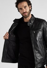 Zwarte leren jas met een ritssluiting aan de voorkant, gewatteerde voering en een zijzak. Heeft een drukknoopkraag en decoratieve stiksels op de schouders.