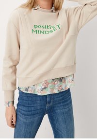 Sweat-shirt beige avec texte vert "positivity", coupe oversize, poignets côtelés, superposé à un chemisier à motifs floraux, porté avec un jean bleu.