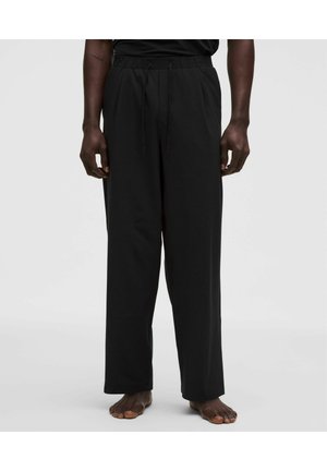 Mann trägt lockere schwarze Hose mit Kordelzug in der Taille, steht barfuß vor einfarbig hellem Hintergrund.