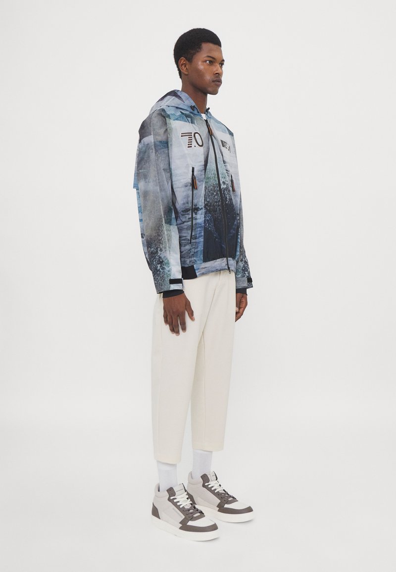 Wasserabweisende Zip-Jacke mit einem blauen und grauen abstrakten Muster, mit Kapuze, seitlichen Taschen und schwarzem Besatz. Getragen mit cremfarbenen Hosen und Sneakers.