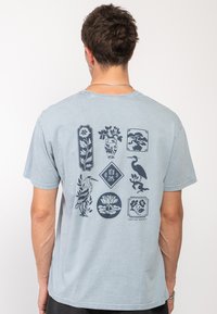 Camiseta de manga corta azul claro con un estampado gráfico en azul oscuro de flores, un pájaro y varias formas decorativas en la parte posterior.