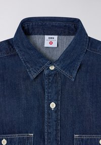Camicia di jeans blu scuro con colletto a punta, chiusura con bottoni bianchi e taschino sul petto. Presenta un'etichetta con marchio e taglia 'M'.