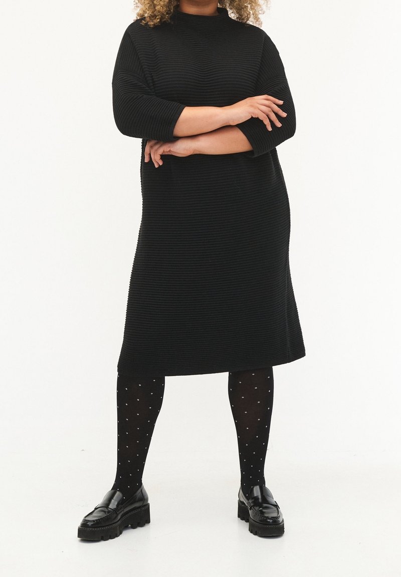 Personne debout, bras croisés, portant une robe noire côtelée, des collants noirs à pois et des mocassins noirs sur un fond blanc.