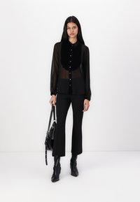 Camisa preta transparente com botões e mangas compridas, acompanhada de calças pretas justas e botas pretas até o tornozelo, segurando uma bolsa preta.
