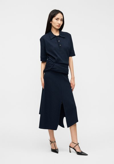 Σετ ρούχων σε σκούρο μπλε, αποτελούμενο από πόλο με κοντά μανίκια, κουμπιά και λεπτομέρεια τύπου wrap, συνδυασμένο με μέσης μήκους φούστα με σχίσιμο στο πλάι.