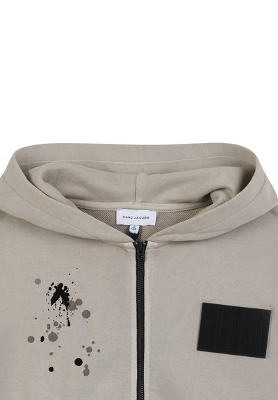 Beige Zip-Hoodie mit glatter Textur, versehen mit einem schwarzen Patch und einem Graffiti-Design in Grau und Schwarz auf der Vorderseite.