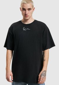 Svart oversized t-skjorte laget av bomull, med korte ermer og rund hals. En hvit skript-logo er plassert på den øvre fronten.