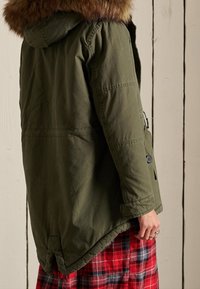Persona che indossa una parka verde oliva con cappuccio in pelliccia, in piedi di lato, abbinata a una gonna a quadri rossi e neri, su uno sfondo di legno.