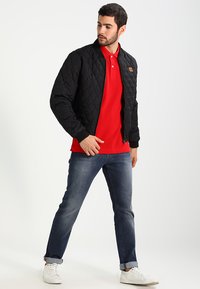 Čierna prešívaná bomber bunda, červená polo košeľa pod ňou, modré džínsy, biele tenisky. Bunda má rebrované manžety a golier. Neformálny outfit.