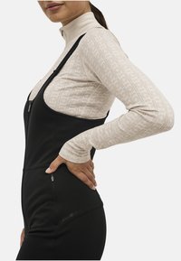 Sort zip-up overall med tekstureret beige langærmet bluse under. Bemærkelsesværdig sidelomme med lynlås og logo detalje på overallen. Glat stof.