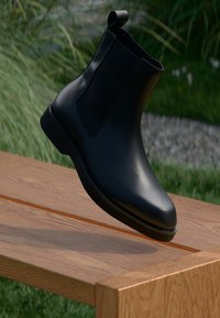 Schwarzer Leder Chelsea-Stiefel mit elastischen Einsätzen, runder Zehenform und niedrigem Absatz, schwebend über einer Holzoberfläche vor einem natürlichen Hintergrund.