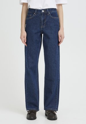 Personne portant un jean droit bleu foncé, un t-shirt blanc et des chaussures noires à lacets, debout devant un fond clair.