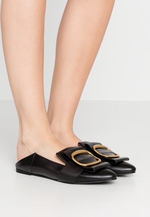 Fødder iført sorte læder slip-on sko med stor guldbeslået spænde på toppen, stående på en glat hvid overflade.