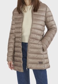 Beige pufferjacka med horisontell quiltad design, dragkedja framtill och sidofickor; har en mjuk textur och en huva. Stylad med en krämfärgad tröja.