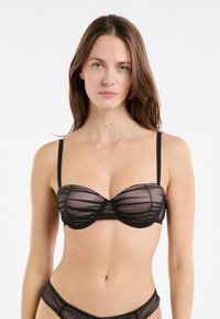 Femme avec de longs cheveux bruns portant un soutien-gorge transparent noir et une lingerie assortie devant un fond blanc uni, faisant face à l'avant.
