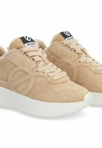 Beige suede sneakers with a textured white sole, rounded shape, lace-up design, and a padded collar. Features a prominent logo on the tongue.