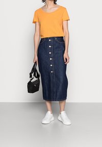 Orange Kurzarmoberteil kombiniert mit einem knielangen dunkelblauen Jeansrock mit einer vorderen Knopfleiste, weißen Sneakers und einer schwarzen Handtasche.