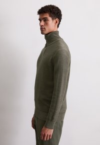 Olivgrüner Pullover mit hohem Kragen, halbem Reißverschluss und gerippten Bündchen. Aus einem strukturierten Strickstoff gefertigt. Figurbetonter Schnitt mit glatter Oberfläche.