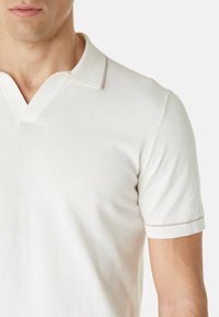 Polo beige clair avec col en V et manches courtes, confectionné en tissu doux avec une bordure contrastante subtile le long du col et des manches.