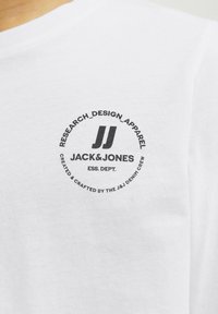 Vit T-shirt i bomull med en cirkulär svart logotyp med texten "JACK&JONES" och "RESEARCH_DESIGN_APPAREL" i fet stil.