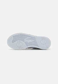 Biała podeszwa sneakerów z wzorem w jodełkę, okrągłymi akcentami i fakturowanymi strefami uchwytu. Wyraziste logo Lacoste wytłoczone z boku.