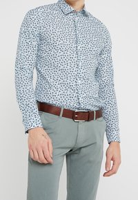 Camicetta azzurro chiaro a maniche lunghe con un motivo floreale, con colletto. Abbinata a pantaloni grigi e una cintura in pelle marrone.