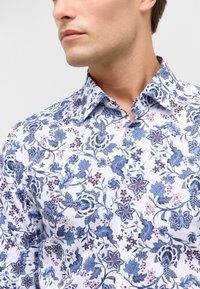 Witte shirt met blauw en paarse bloemmotieven, met een kraag en knoopsluiting. Gestructureerde stof met ingewikkelde ontwerpelementen.