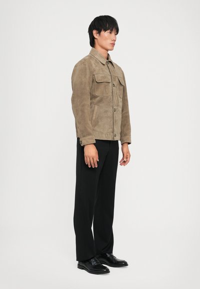 Brown suede jacket with a collar, two front pockets, and button closure, paired with black trousers and black shoes. Simple and structured design.