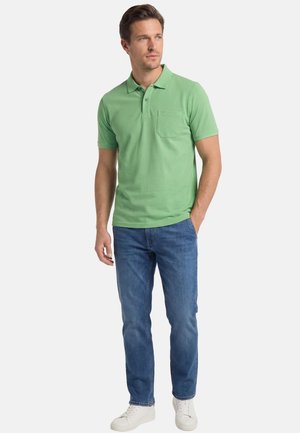 Mann trägt ein hellgrünes Kurzarm-Poloshirt, blaue Jeans und weiße Turnschuhe, steht mit einer Hand in der Tasche.