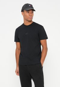 Hombre con una gorra negra de Tommy Jeans, una camiseta negra de cuello redondo con un pequeño logo y pantalones negros, de pie con una mano en el bolsillo, mirando a la derecha.